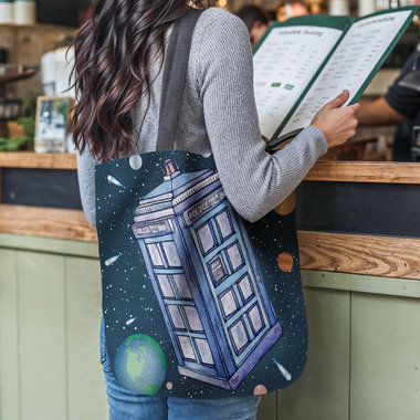 Police call box floating in the solar system tote bag hanging on the shoulder of a woman at a restaurant counter.